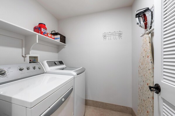 A laundry room with a front-loading washer and dryer, a shelf with laundry supplies, and a drying rack on the wall. - Ocean Front With Stunning Views! Halii Kai 13E (Waikoloa Village)