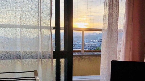 A room with a bed, a TV on a small table, a window with sheer curtains, and a view of buildings and a sunset. - Ichijyo-IVY Semi double (Kyoto)