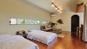 A modern bedroom with two beds, a wooden floor, a sofa, a small table, and a window with a view of greenery. - IMAGINE OKINAWA (Nakijin)