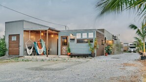 A small, single-story building with a corrugated metal exterior and a wooden door. There are two hammocks hanging outside, a bench, and several potted plants. - IMAGINE OKINAWA (Nakijin)