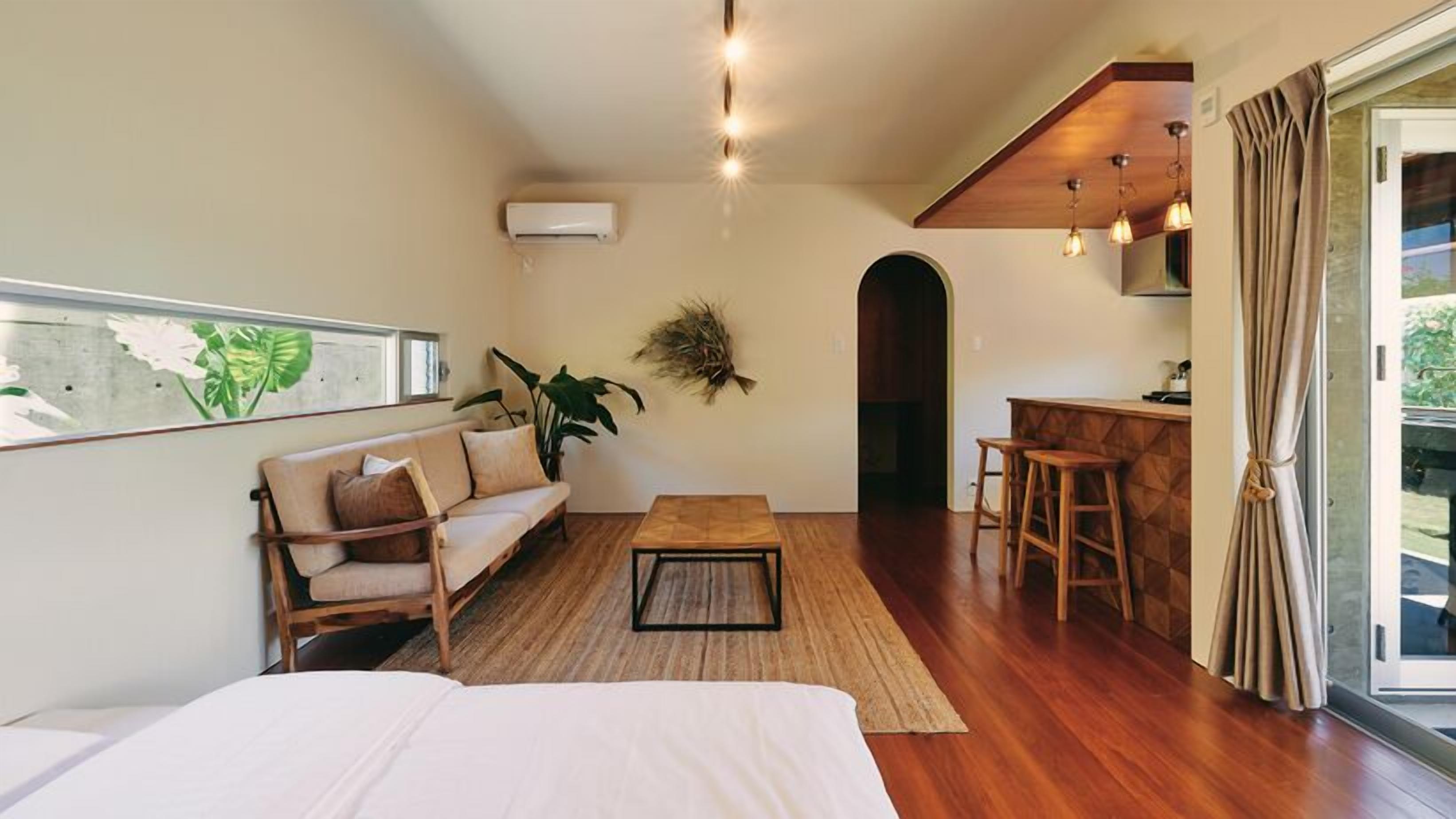 A room with a bed, a sofa, a coffee table, a small kitchen area with stools, a hanging plant, and a window with a view.