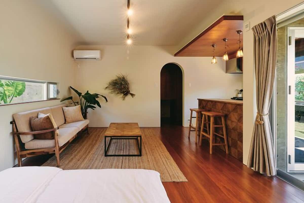 A room with a bed, a sofa, a coffee table, a small kitchen area with stools, a hanging plant, and a window with a view.