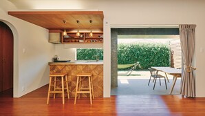 A modern kitchen with wooden accents, a dining area with a view of a green hedge, and a large window. - IMAGINE OKINAWA (Nakijin)