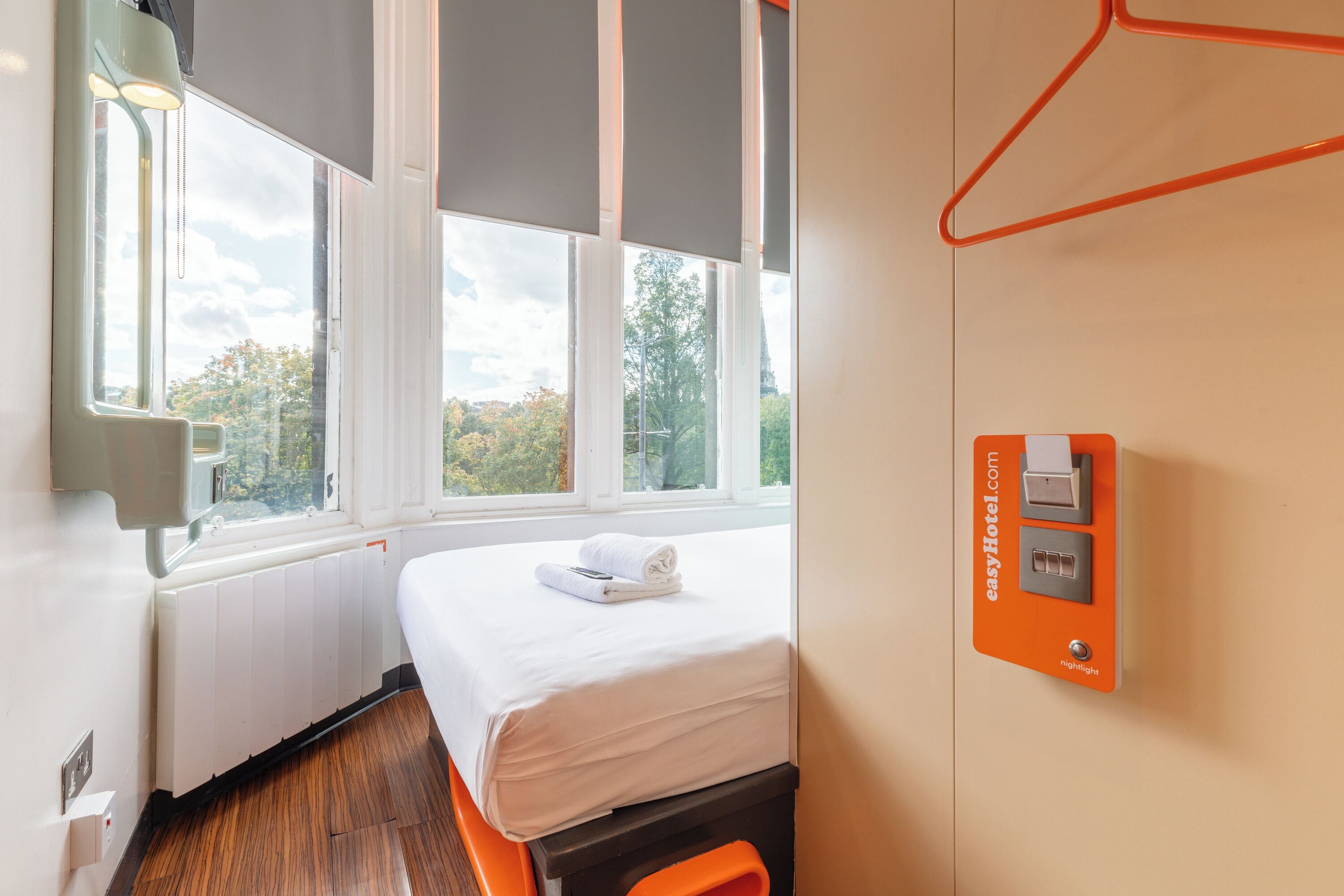 A hotel room with a bed, a window with a view of trees, a radiator, and a control panel.