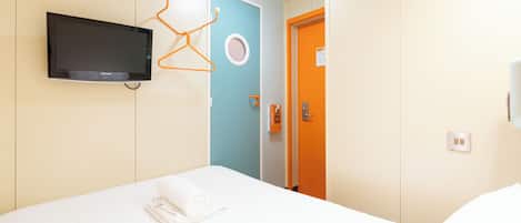 A hotel room with a bed, a television mounted on the wall, a door with a circular window, and a towel on the bed.