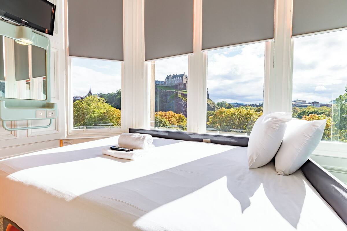 A hotel room with a large bed, a TV, and a view of a castle through the windows.