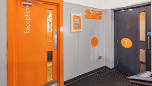 Una zona d'entrada amb una porta taronja brillant amb la inscripció 'Reception' i un senyal que indica els números de les habitacions de 1.1 a 1.9.