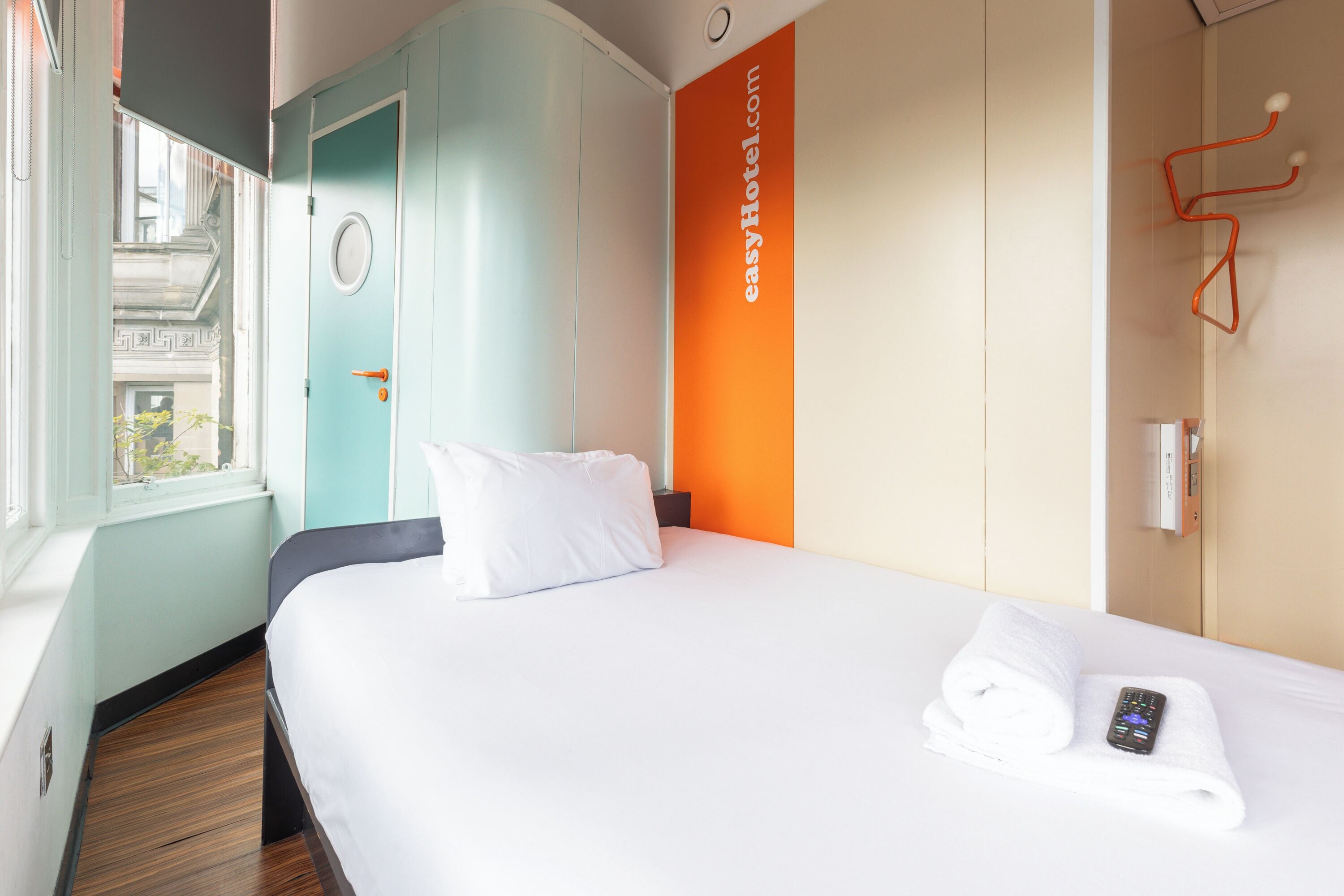 A hotel room with a bed, a window, a door, and a wall-mounted control panel.