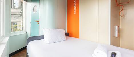 A hotel room with a bed, a window, a door, and a wall-mounted control panel.