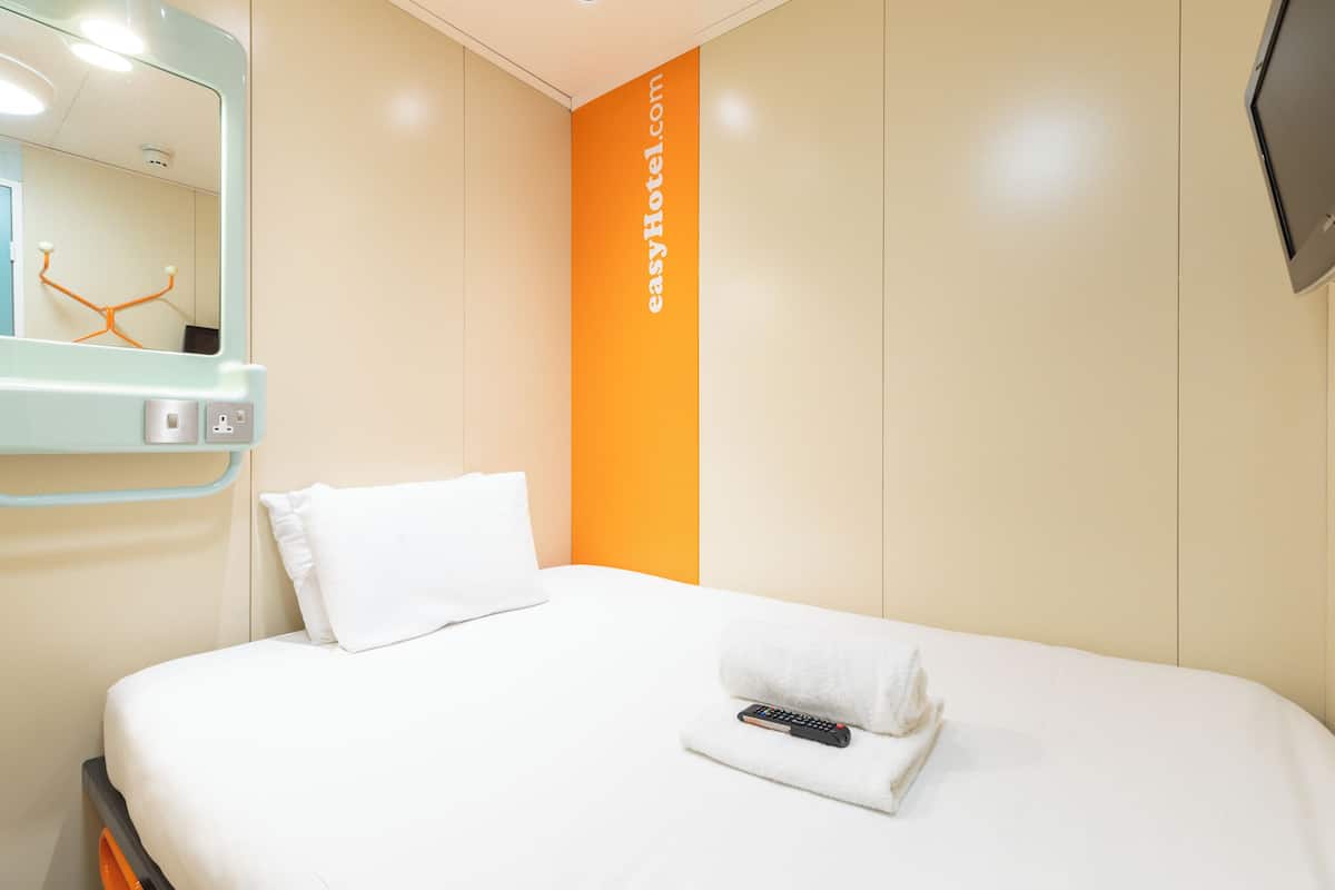A hotel room with a bed, a white pillow, a remote control, and a wall with a vertical orange stripe.