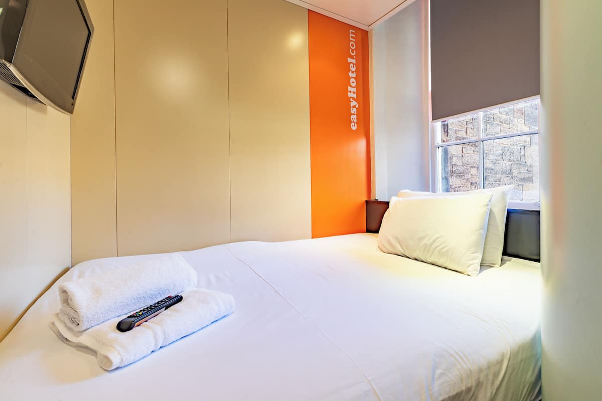 A hotel room with a bed, a remote control, and a window with a view of a stone wall.
