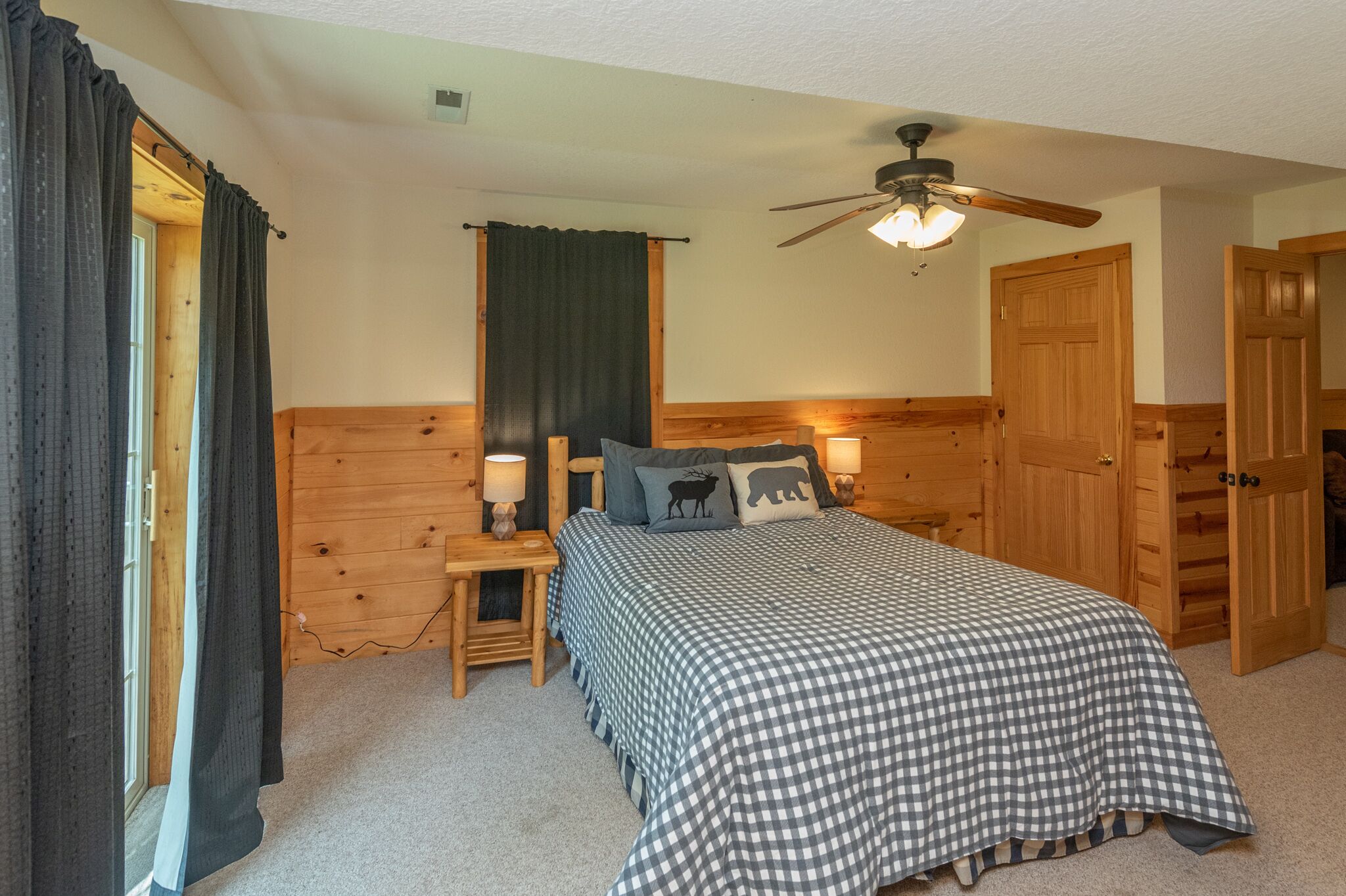 A bedroom with a checkered bedspread, wooden headboard, two bedside lamps, a ceiling fan, and a door with a wooden panel.