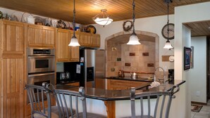 A kitchen with wooden cabinets, stainless steel appliances, a central island with chairs, pendant lighting, and a tiled backsplash.