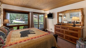 A bedroom with a large bed, a wooden dresser, a television, and a balcony with a view of the outdoors.