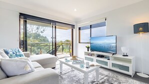 Ein modernes Wohnzimmer mit großem Fernseher, weißen Möbeln und einem Balkon mit Meerblick.