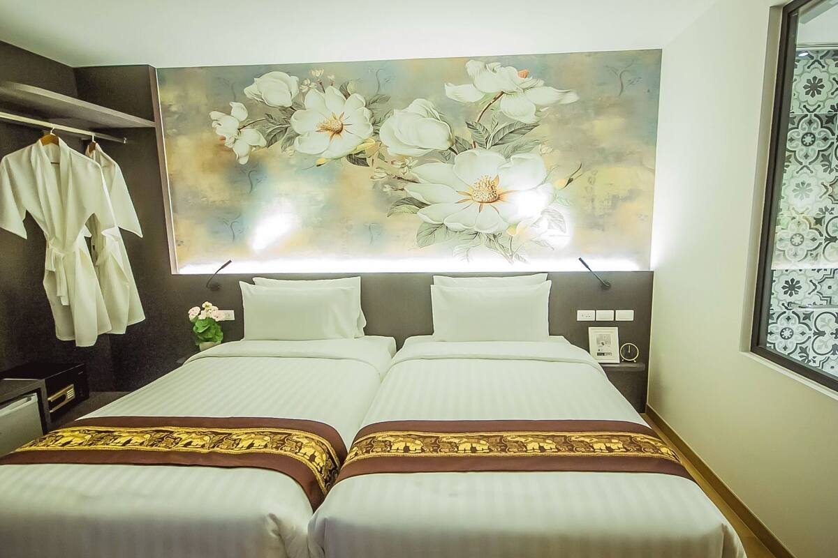 Une chambre d’hôtel avec deux lits, une grande œuvre d’art florale accrochée au mur, un porte-peignoir et une fenêtre donnant sur un paysage aux motifs variés.