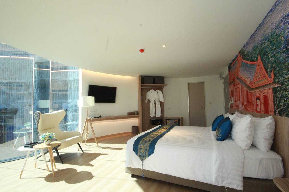 Une chambre d’hôtel moderne dotée d’un grand lit, d’un coin salon avec un fauteuil et une table, d’une armoire et d’une grande fenêtre donnant sur la ville.
