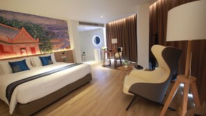 Hotellihuone, jossa on suuri sänky, oleskelualue tuolilla ja pöydällä, seinään kiinnitetty valaisin ja koristeellinen seinävaate.