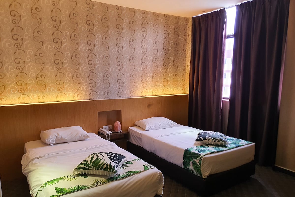 Habitación de hotel con dos camas, papel tapiz estampado, cabecera de madera, teléfono y ventana con cortinas.