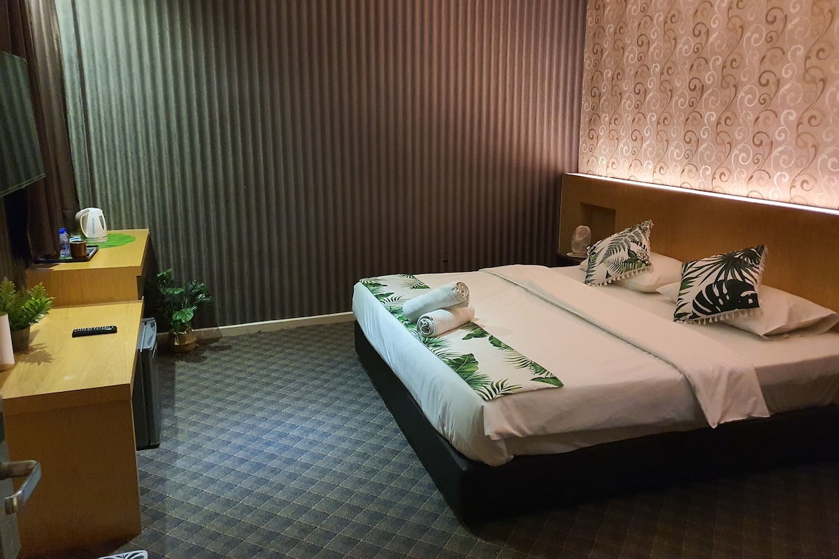 Una habitación de hotel con cama, escritorio con hervidor, control remoto y una pequeña planta.