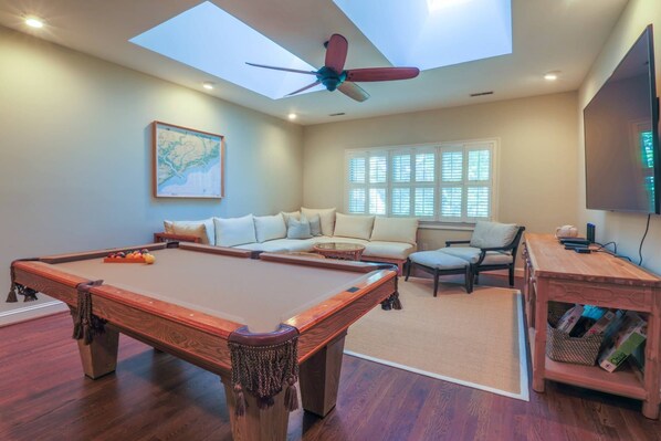 A room with a pool table, a sofa, a chair, a television, and a ceiling fan.