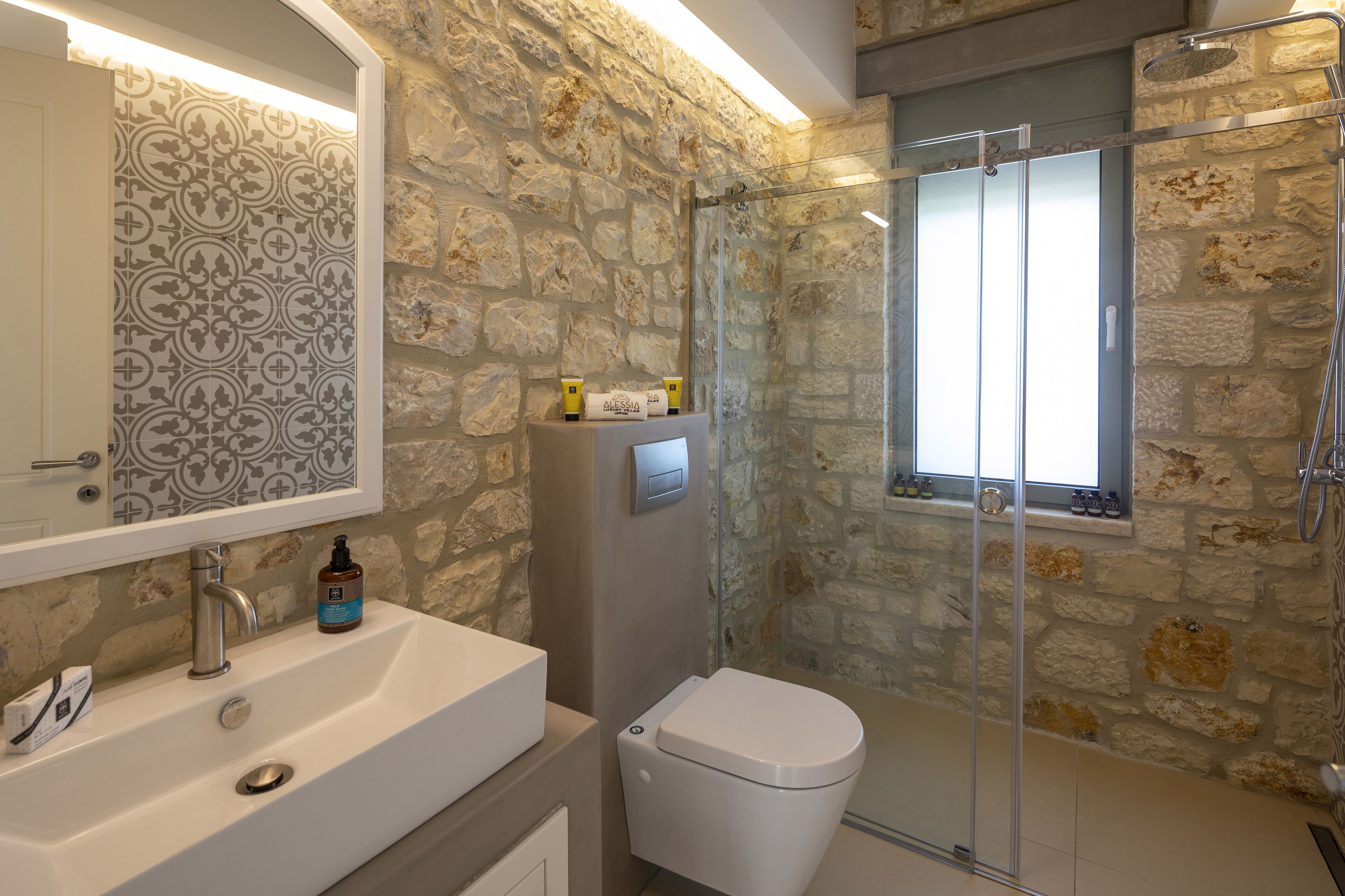 A bathroom with a stone wall, a glass shower enclosure, a white sink, and a toilet.