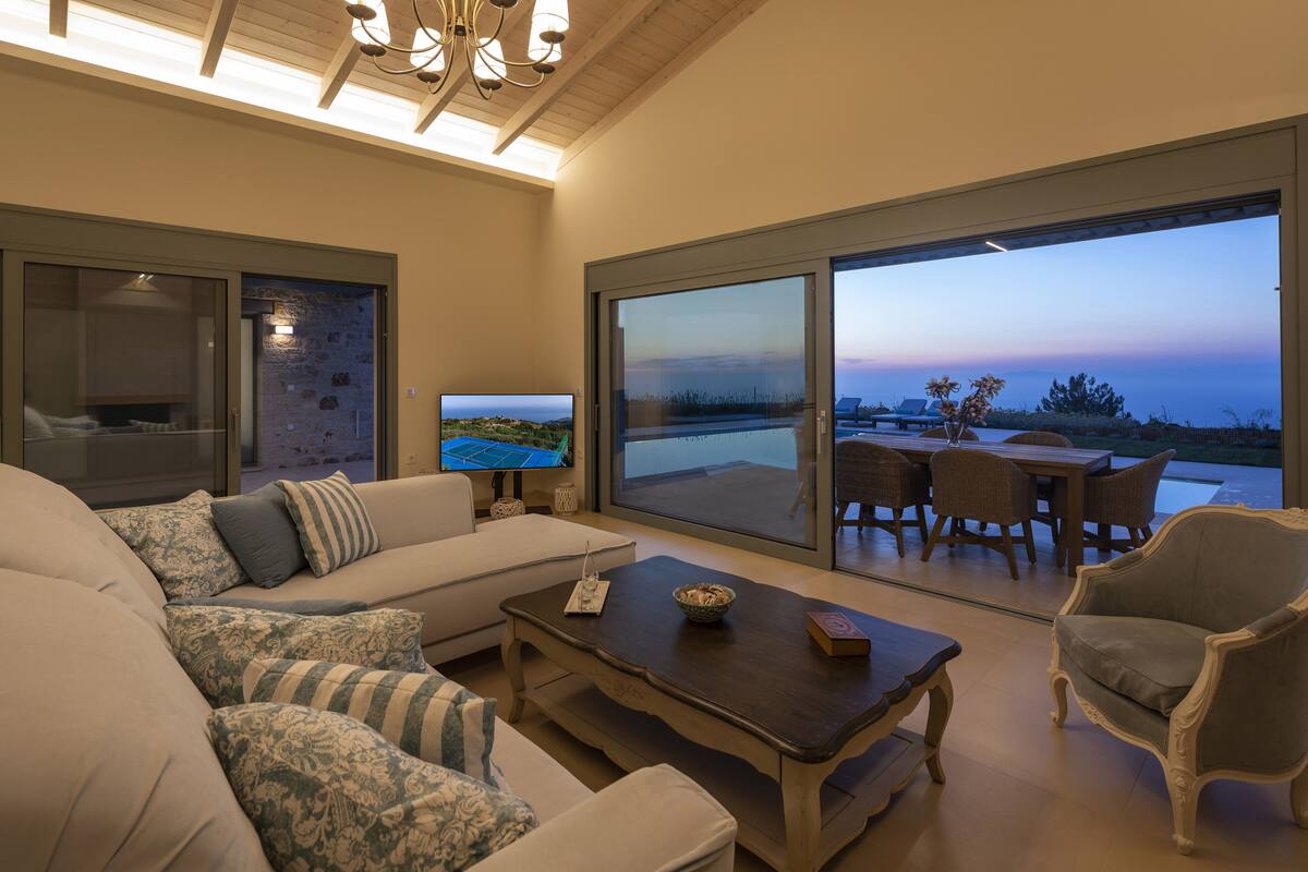 A modern living room with a large sofa, a coffee table, and a TV. There is a sliding glass door leading to an outdoor patio with a dining table and chairs.