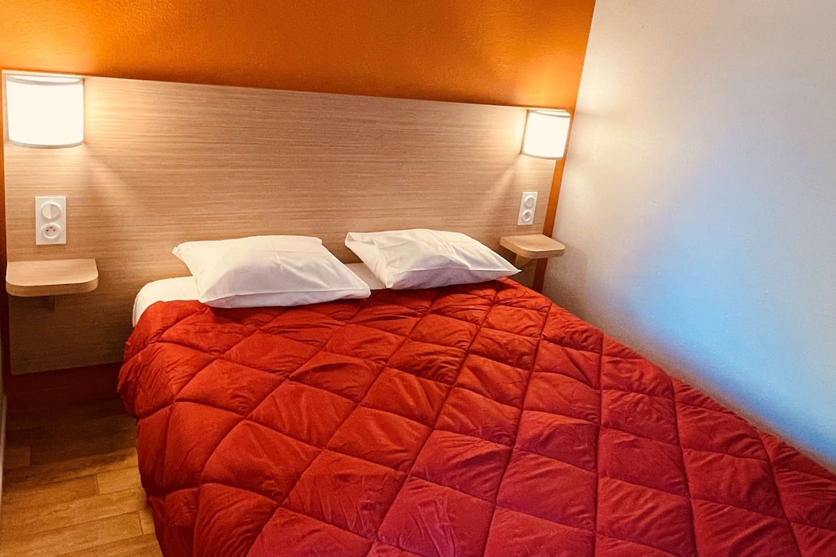 Quarto de hotel com colcha de cama acolchoada vermelha, cabeceira de madeira, duas mesas de cabeceira com abajures e piso de madeira.