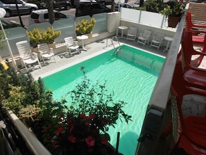 A rooftop pool with white chairs and tables, surrounded by potted plants and a view of parked cars. - Hotel Arno (Rimini)