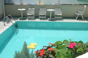 Une piscine avec un escalier en métal, entourée de chaises longues blanches et de petites tables rondes.