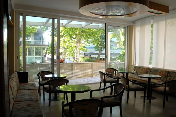 A modern indoor seating area with a sofa, chairs, and tables, large windows with a view of greenery and buildings outside. - Hotel Arno (Rimini)