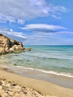 A coastal beach with clear water, rocky cliffs, and a sandy shore.