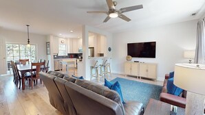 A spacious living room with a leather sofa, a dining area with wooden chairs, a kitchen with white cabinets, and a ceiling fan.