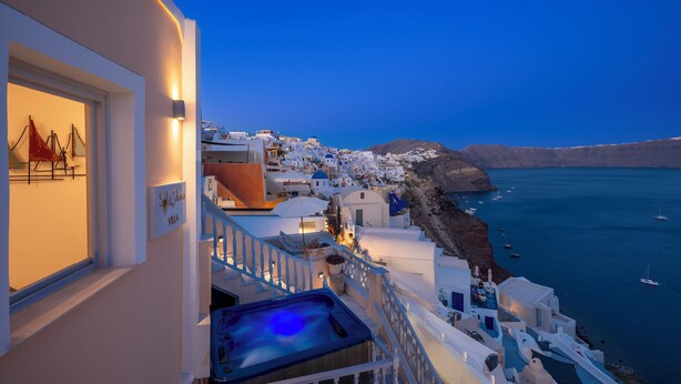 A coastal hotel with white buildings, a balcony with a hot tub, and a view of the sea and boats. - Sole d'oro Sunset & Caldera View Grand Suite (Santorini)
