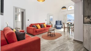 A spacious living room with a red sofa, a coffee table, and a dining area with a table and chairs. There is a kitchen area with a stove and cabinets. - Sole d'oro Sunset & Caldera View Grand Suite (Santorini)