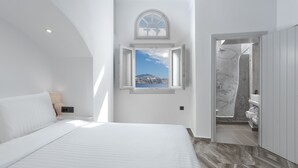 A bedroom with a bed, a nightstand, a window with an open view, and a bathroom visible through an open door. - Sole d'oro Sunset & Caldera View Grand Suite (Santorini)