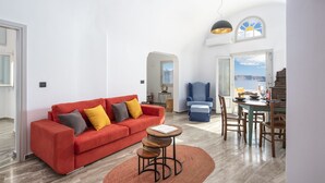 A spacious living area with a red sofa, a dining table with chairs, and a view of the sea through a window. - Sole d'oro Sunset & Caldera View Grand Suite (Santorini)
