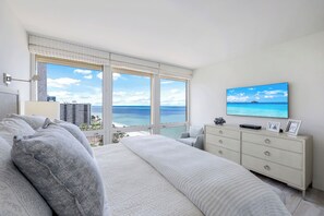 A bedroom with a large bed, a dresser, a TV, and a view of the ocean.