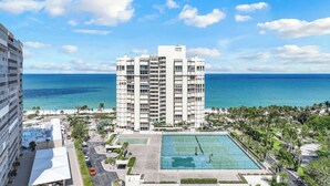 A high-rise hotel with a tennis court and pool area, overlooking a beach and ocean.