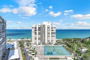 A high-rise hotel with a tennis court and pool area, overlooking a beach and ocean.