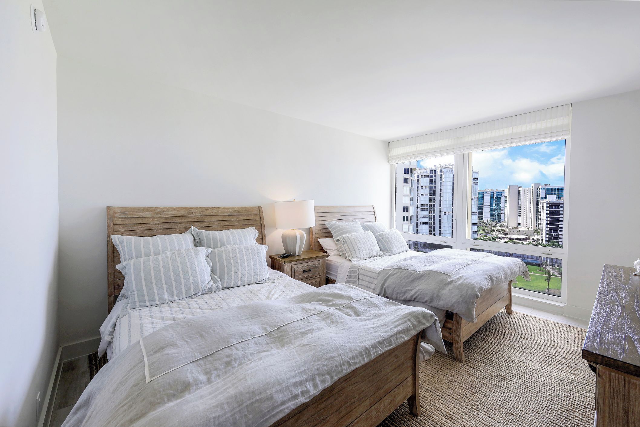 A hotel room with two beds, a nightstand, a lamp, and a view of the cityscape.