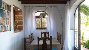 Un área de comedor con una mesa y sillas de madera, pared de ladrillo con un mural colorido, una ventana con cortinas blancas y vista a la vegetación exterior.