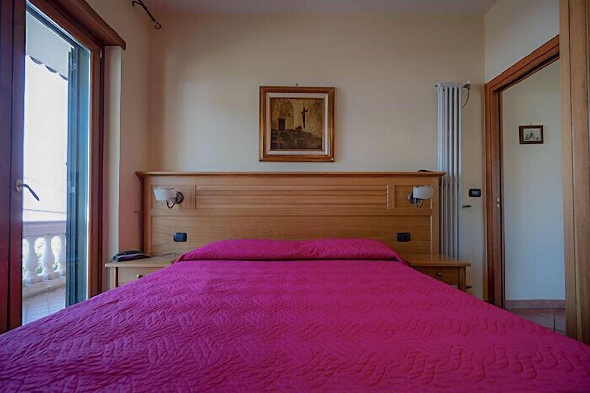 Een slaapkamer met een groot bed, een nachtkastje, een houten hoofdbord, een ingelijste foto aan de muur en een raam met gordijnen.