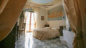 Une chambre à coucher avec un lit, une chaise, un bureau, une fenêtre avec des rideaux, un tableau au mur et un sol carrelé.