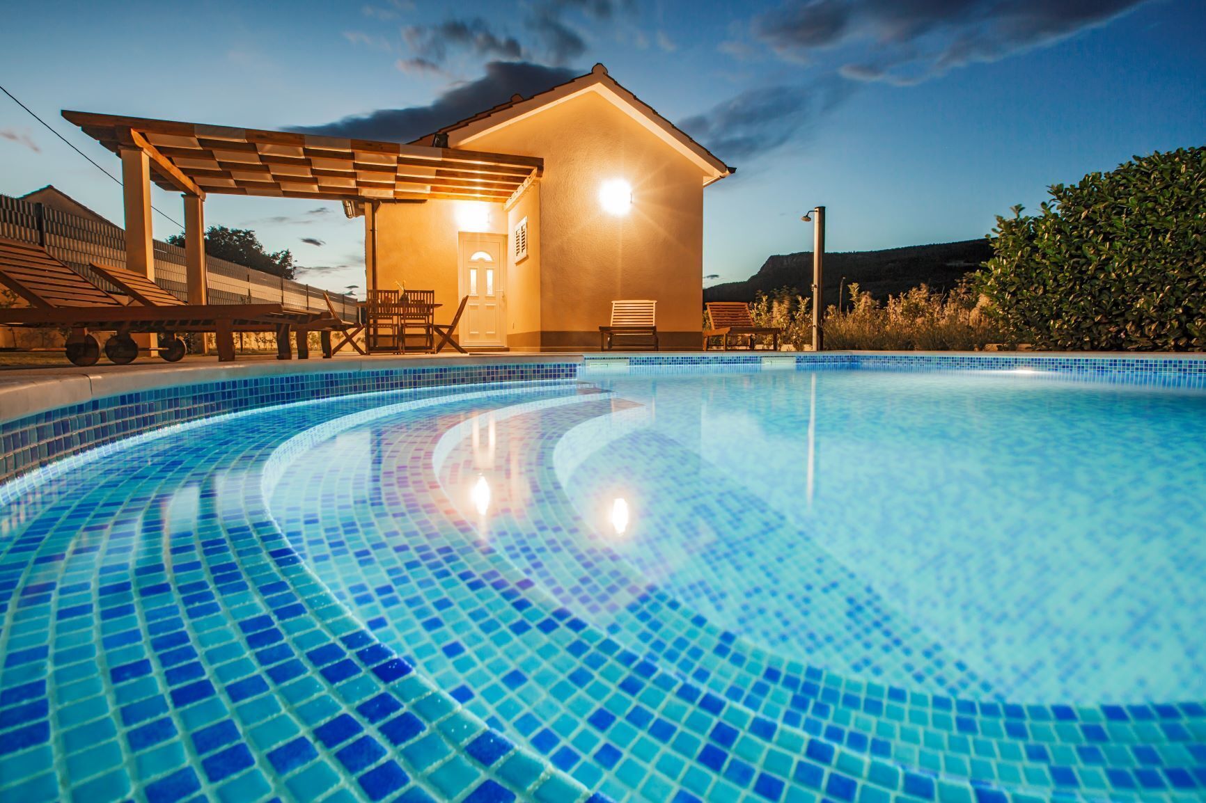 A swimming pool with blue mosaic tiles, a wooden pergola, and a small building with a light on one side.