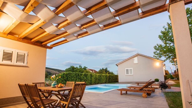Un patio exterior con piscina, mobiliario de madera y una casa al fondo.