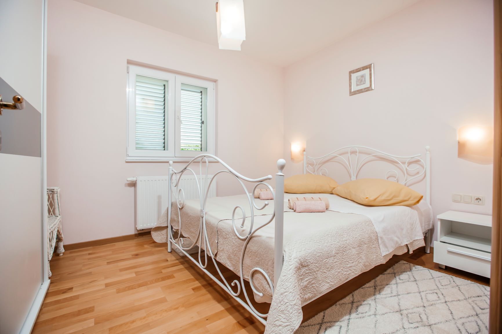 Ein Schlafzimmer mit weißem Metallbettgestell, einem weißen Heizkörper, einem Fenster mit Jalousien und einem Nachttisch mit Lampe.