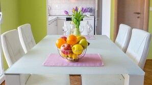 Mesa de jantar com uma fruteira, cadeiras brancas e uma cozinha ao fundo.