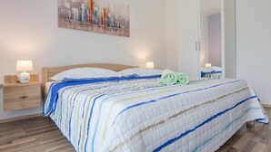 Uma cama bem arrumada com colcha listrada, uma mesinha de cabeceira com abajur e uma pintura com vista para a cidade na parede.