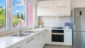 Uma cozinha moderna com armários brancos, eletrodomésticos de aço inoxidável, uma janela com vista para o verde e um pequeno vaso de planta no balcão.